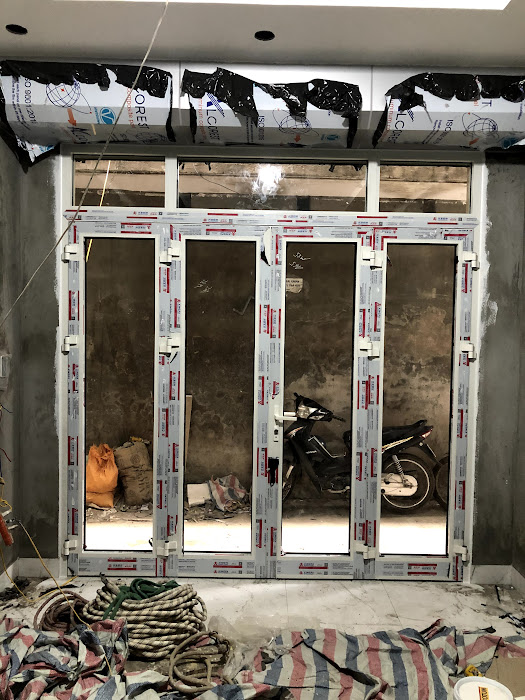 Cửa nhôm kính bốn cánh đang lắp đặt trong nhà xây dựng dở, nhìn ra sân có xe máy
